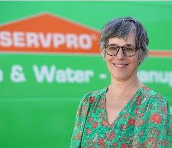 Middle-aged woman with short, partially gray hair wearing glasses and a floral pattern shirt, standing by a SERVPRO truck